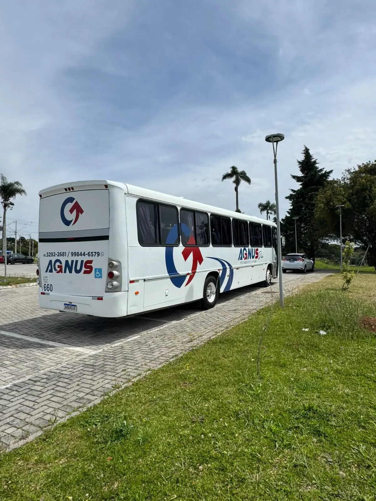 Traseira e lateral de um ônibus da Agnus, com o símbolo da marca, ao lado de uma área verde.
